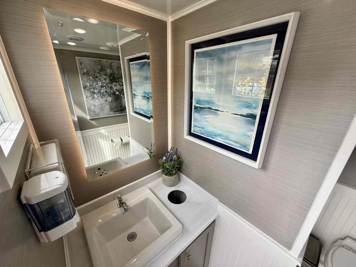 Interior of a luxury restroom trailer with a flushing toilet, sink, mirror, and lighting.