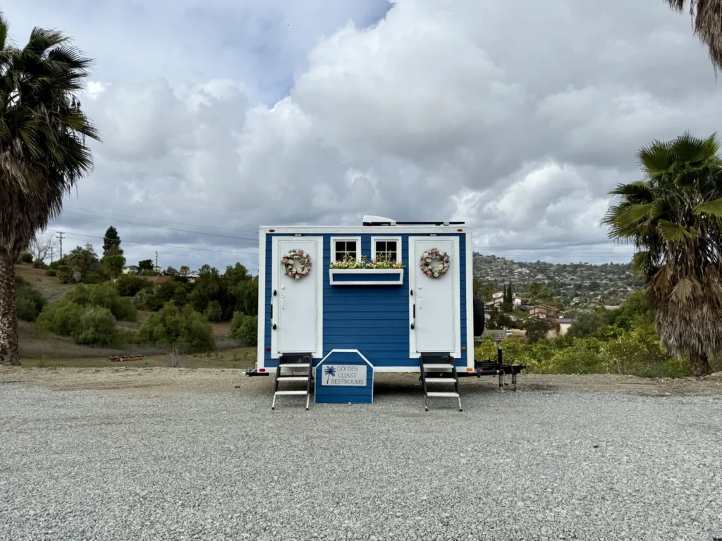 Golden Coast Restrooms blue cottage restroom trailer with wreaths and flower boxes set up on a gravel lot with inland North County hills behind it, representing restroom trailer rental in Escondido, CA.