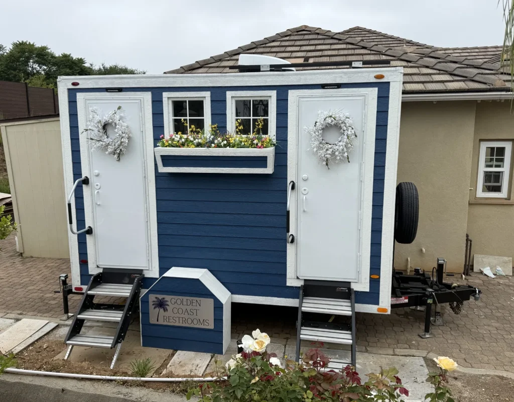 Golden Coast Restrooms 2-Stall Blue Cottage restroom trailer with flower boxes and a wreath set up beside a home in San Diego, CA.