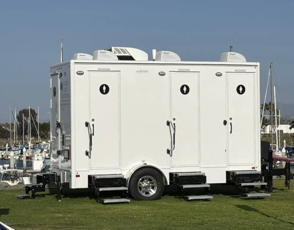 White Golden Coast Restrooms luxury restroom trailer with separate stall doors parked on a lawn beside a harbor for a public event in coastal San Diego County.