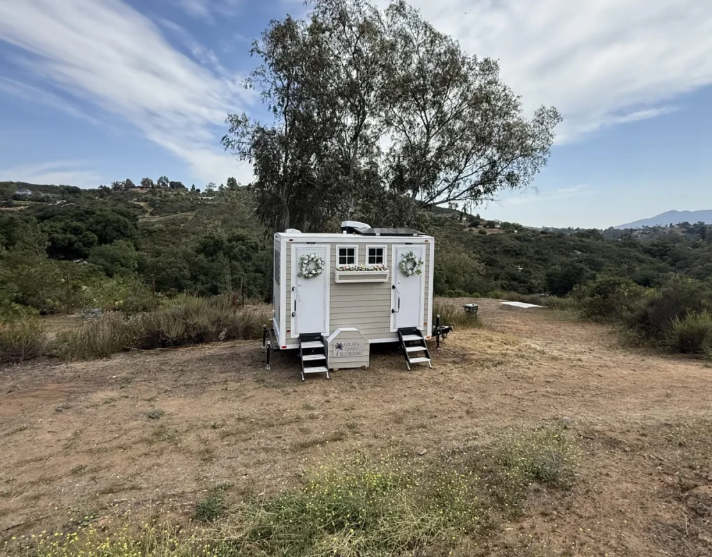 Restroom trailer parked in an open field beside a large tree on a rural property, the kind of ranch event site served in Escondido, CA.