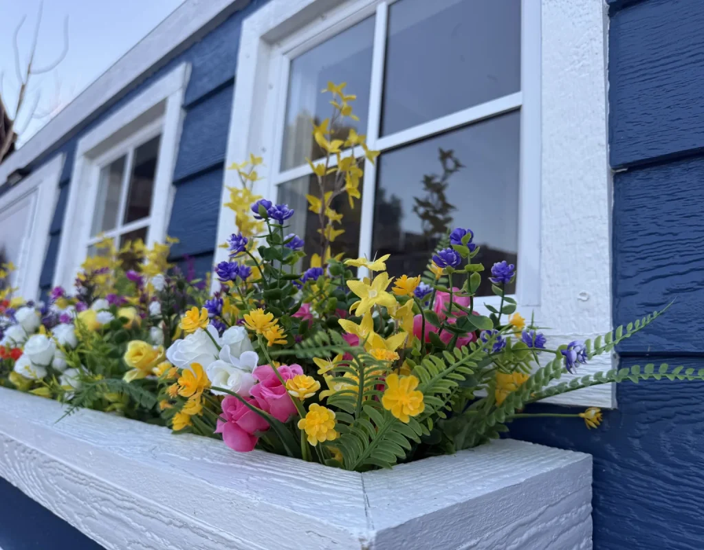 Flower box filled with yellow, pink, and white flowers under a window on a blue cottage restroom trailer prepared for a Carlsbad, CA wedding.